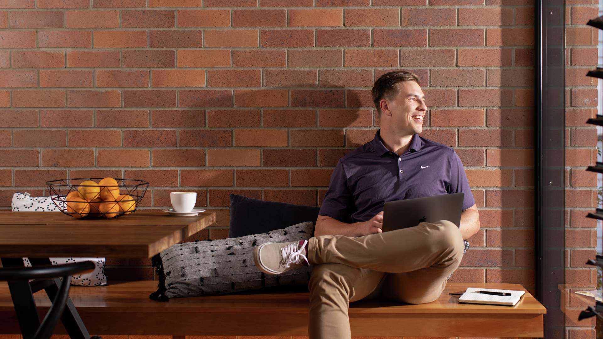 A student sits at home on a bench with a computer on his lap.