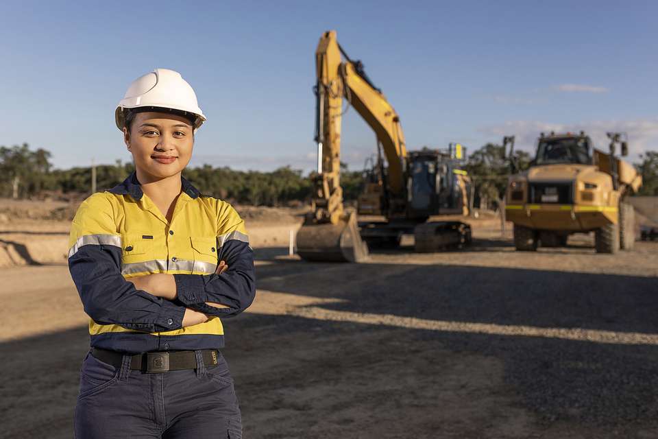 Engineering worker in hi vis and a hard hat stands in front of a worksite and machinery.