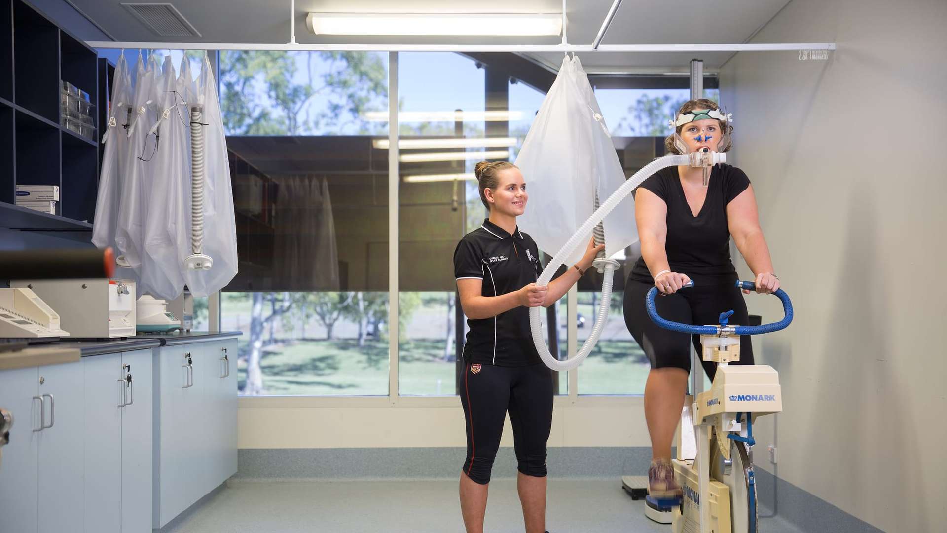 A person stands beside another individual who is using a stationary exercise bike, holding a tube connected to the equipment