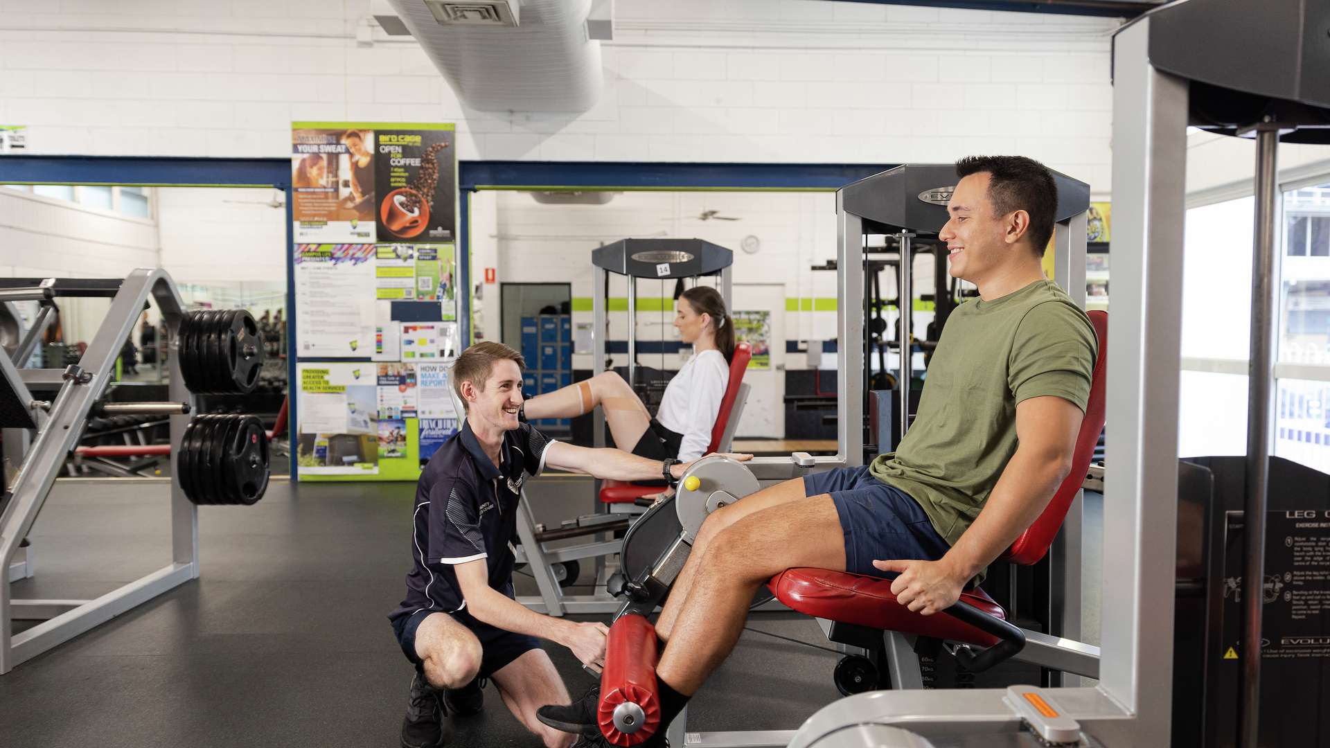 A student using some gym equipment is supervised by a trainer.