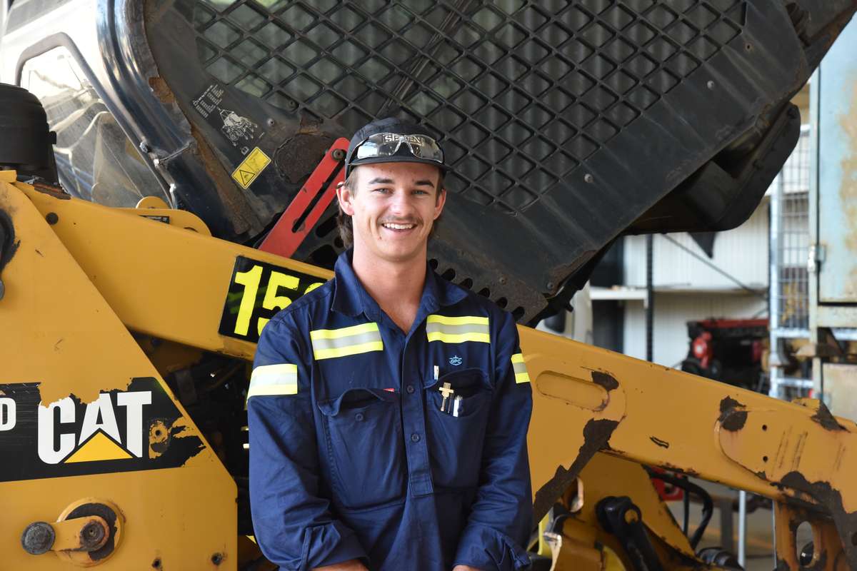 A young man in a blue and yellow hi-visibility workshirt standing in front of a heavy vehicle