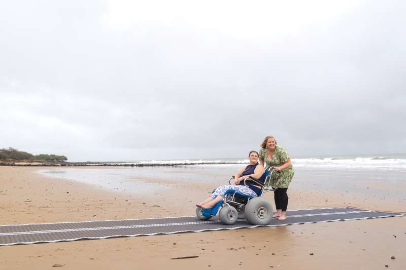 Student helping another student in a wheelchair down the beach smiling at the ocean.