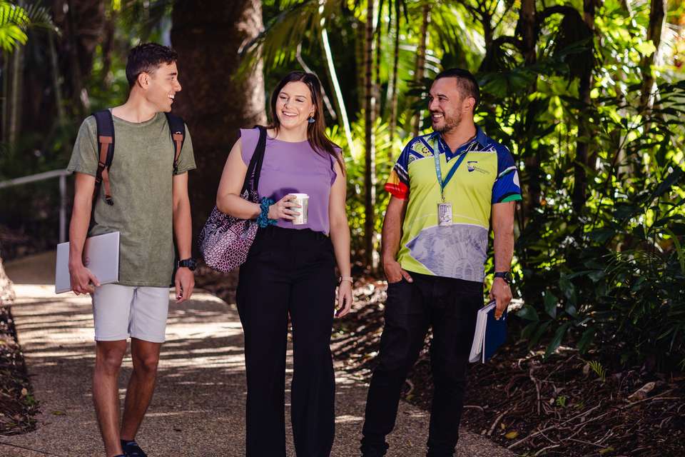 Three CQU students walking along path smiling, one holds a drink, two holding notepads