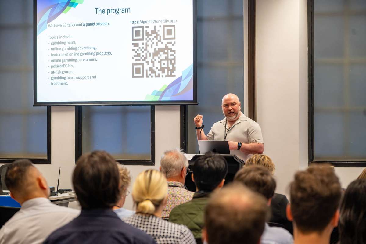 Prof Alex Russell stands at a lectern in front of a big screen, a large seated crowd look towards him.