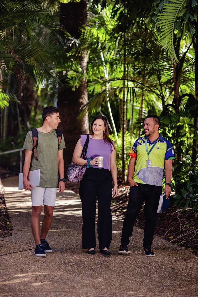 Three CQU students walking along path smiling, one holds a drink, two holding notepads