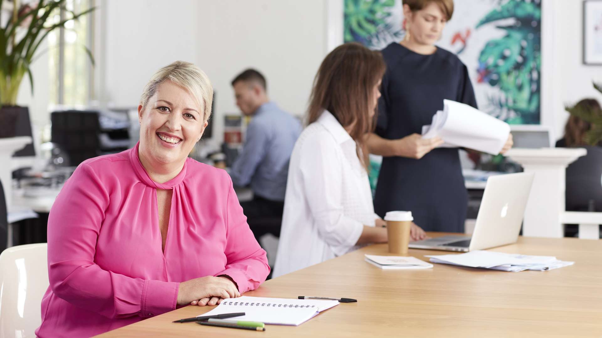 Student sits at a desk in open workspace with students in the background talking to the lecturer.