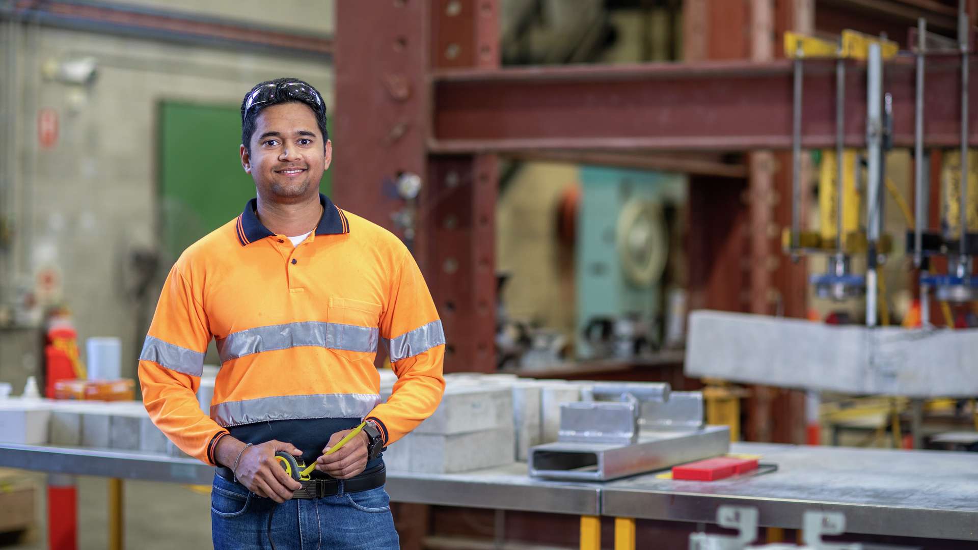 Civil Engineering student stands in engineering work shop with building materials on bench behind him.