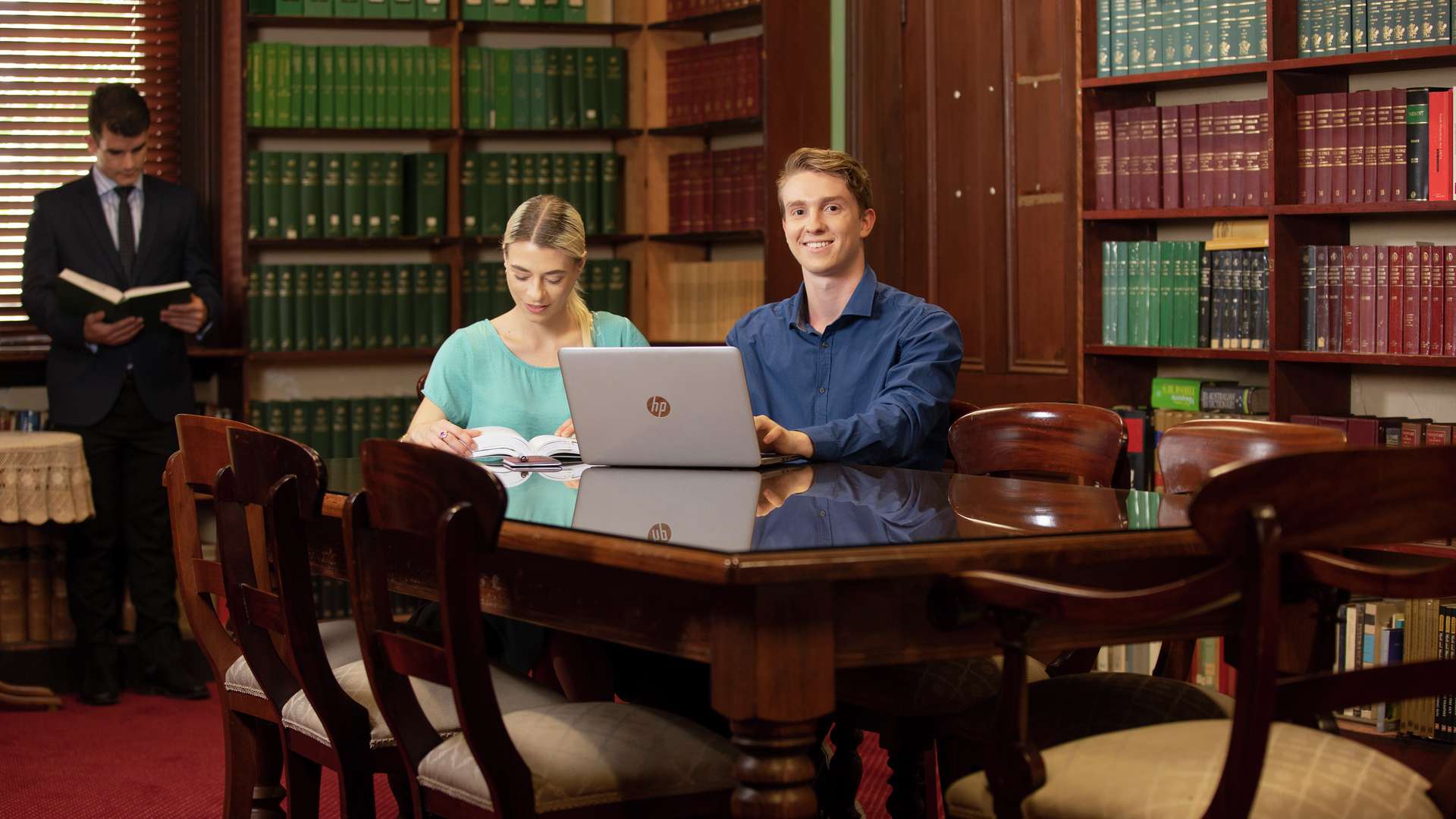 Two law students seated at a table, one using a laptop, another reading a book, while a third student reads in the background near shelves with legal books.