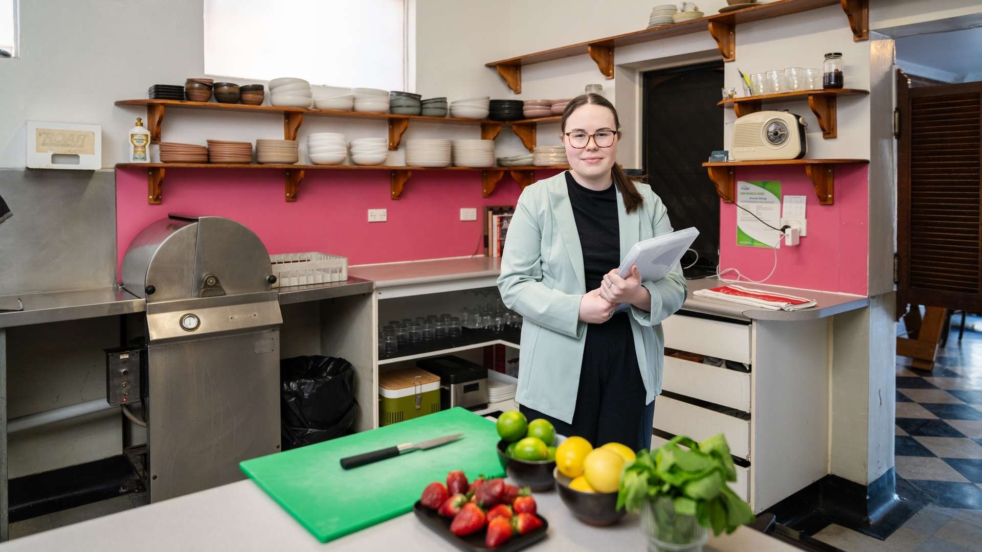Student standing in working kitchen holding clipboard with fruit and cutting board on bench in foreground.