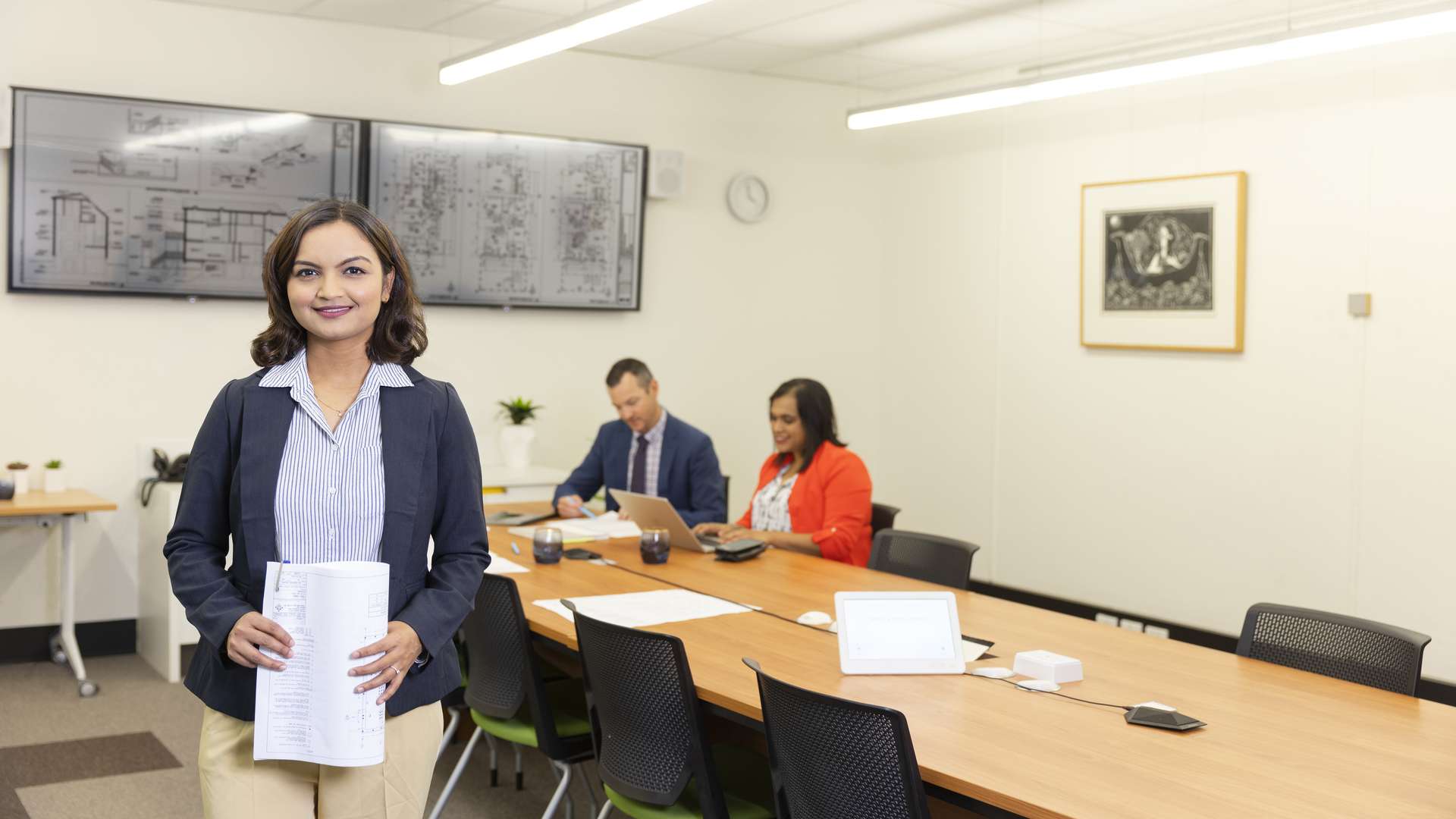 Person in business attire holding paperwork with two colleagues working at board table in background
