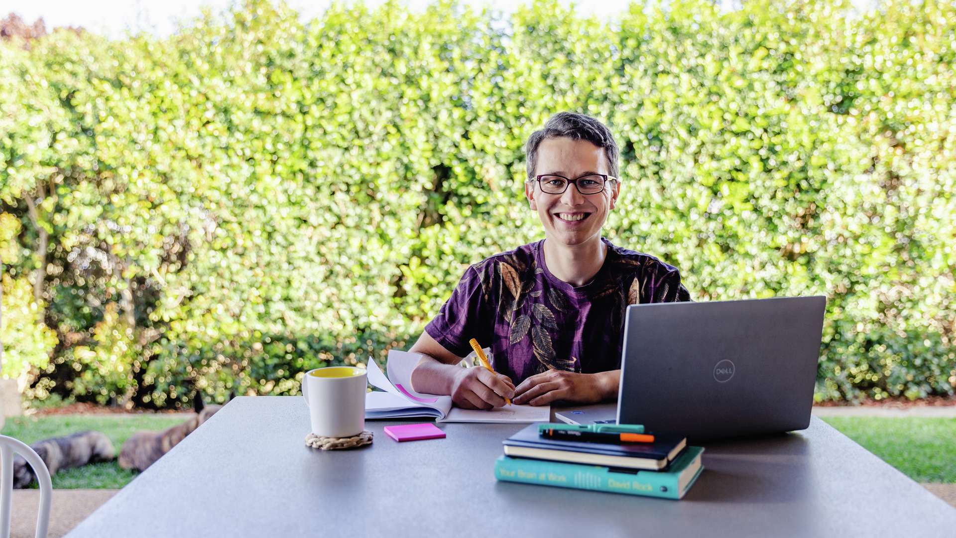 Student sitting at table outside with workbooks and laptop taking notes in notebook.