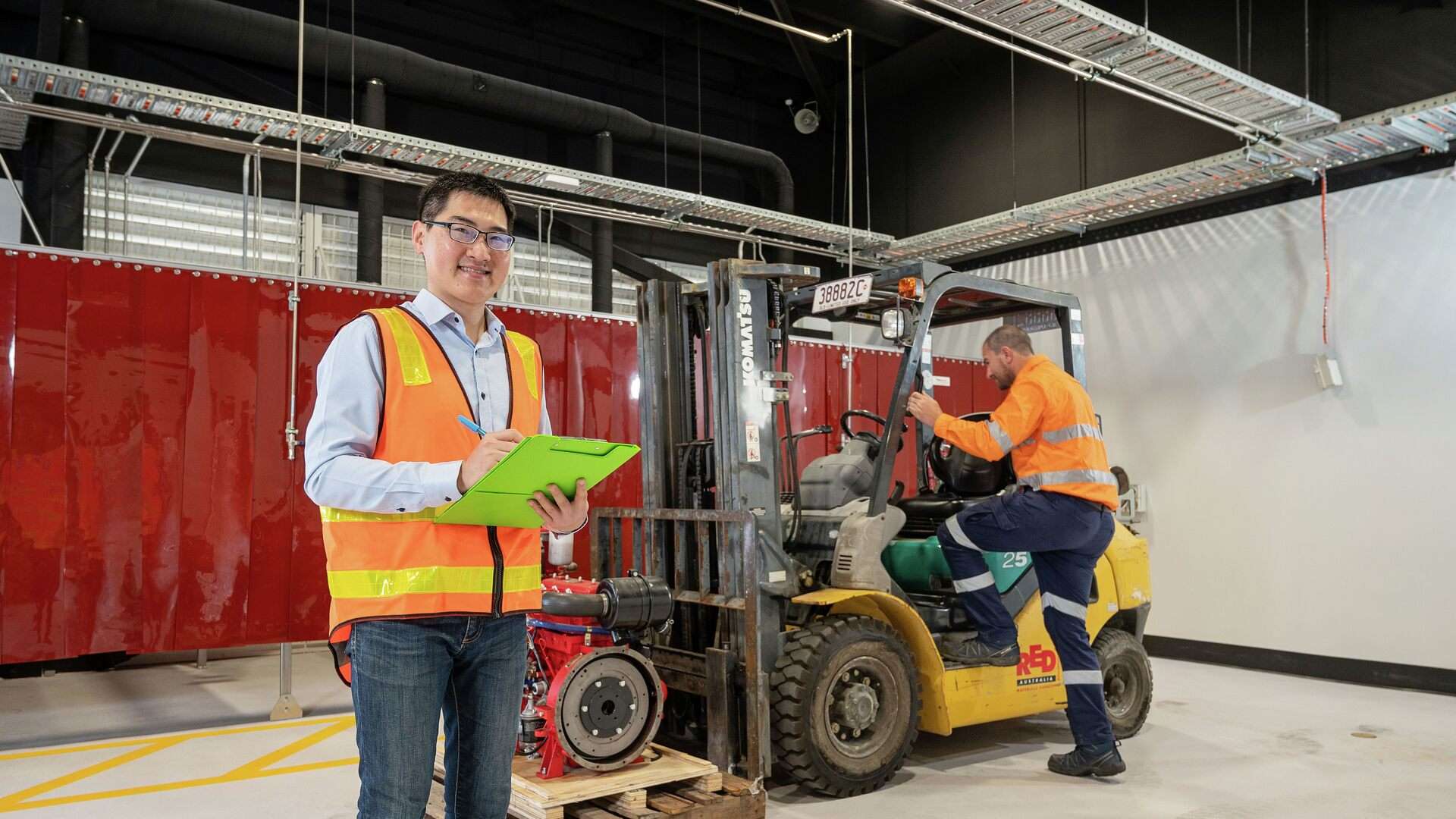 A student wearing a high vis vest holds a clipboard while a person in high vis workwear climbs into a forklift in the background.