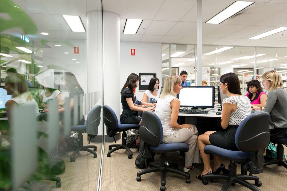A group of eight CQU students in the library computer lab sitting on office chairs in front of a circular desk with computers. They are using the computers to study.