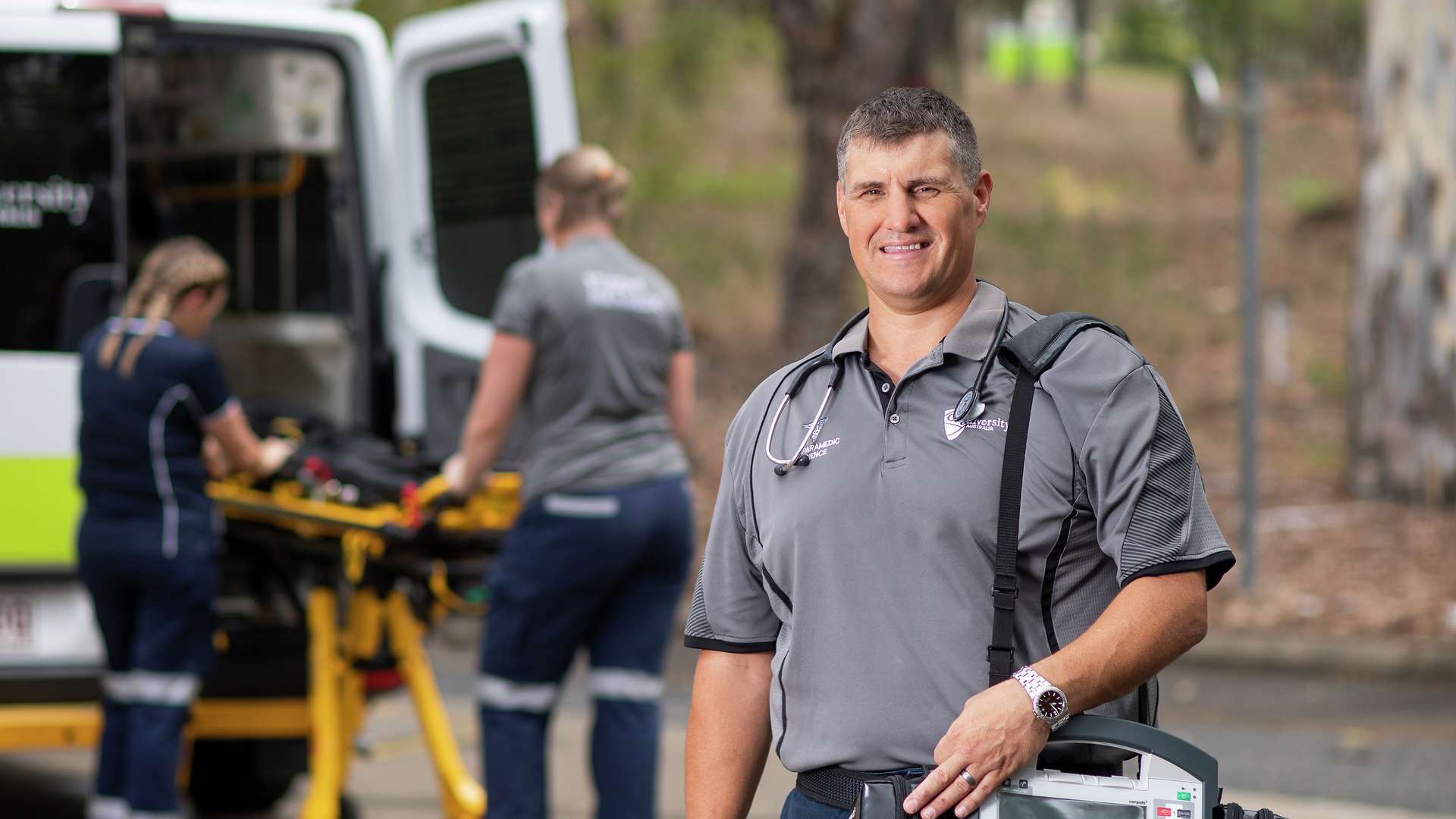 A paramedic stands in front of an ambulance, where two colleagues are handling a gurney.