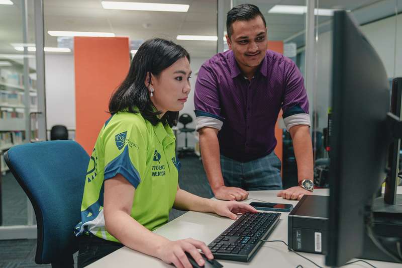 There are two CQU students in front of a computer in the library. One is a CQU student mentor who is sitting on a chair in front of the computer. The other student is standing next to them watching the computer screen.