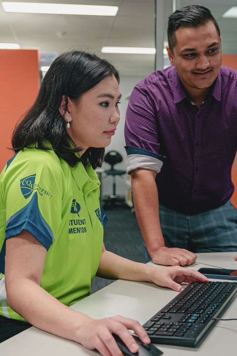 There are two CQU students in front of a computer in the library. One is a CQU student mentor who is sitting on a chair in front of the computer. The other student is standing next to them watching the computer screen.