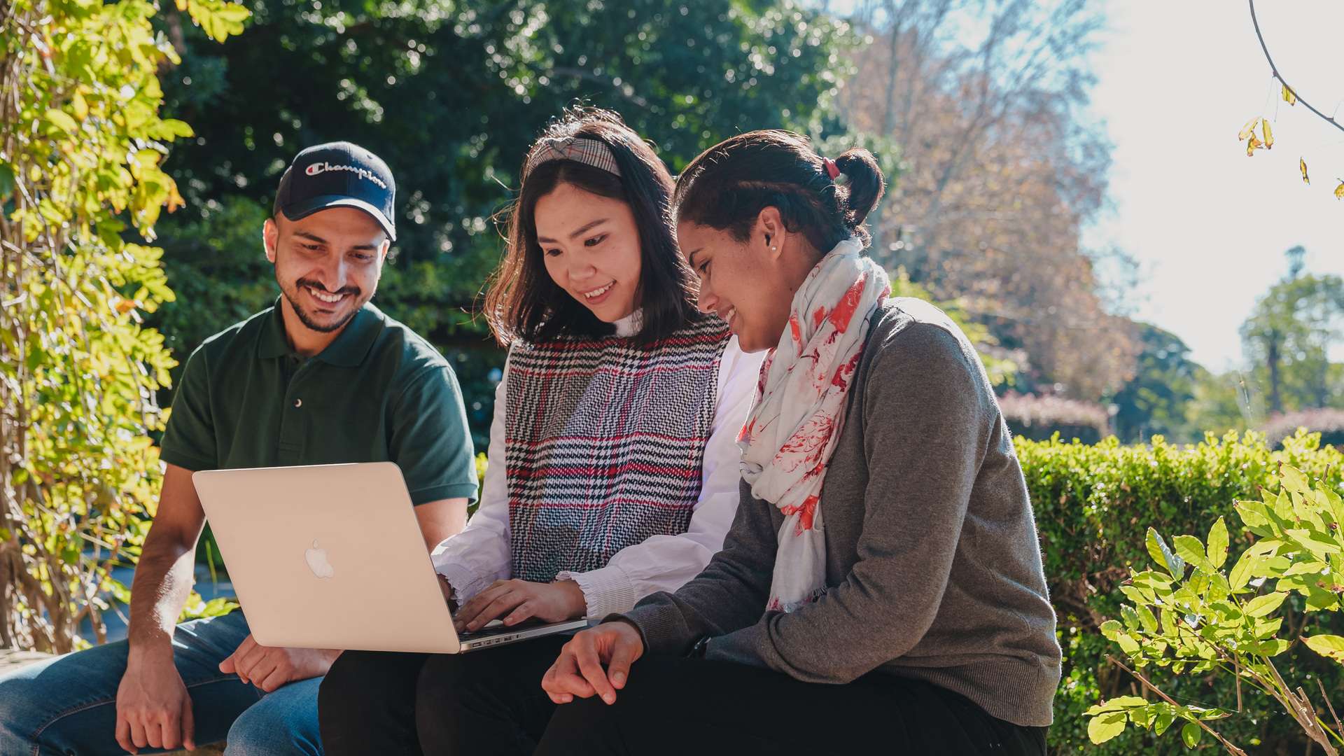 Three students sitting outside, the outer two students focusing on the centre student using their laptop