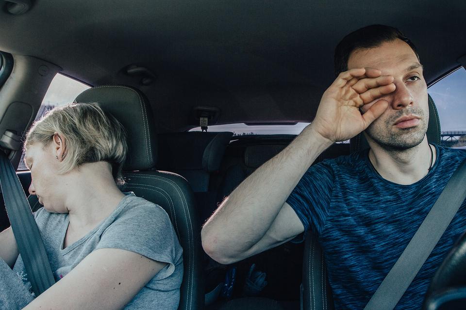 Passenger in a car sleeping as the driver looks visibly tired rubbing their eye