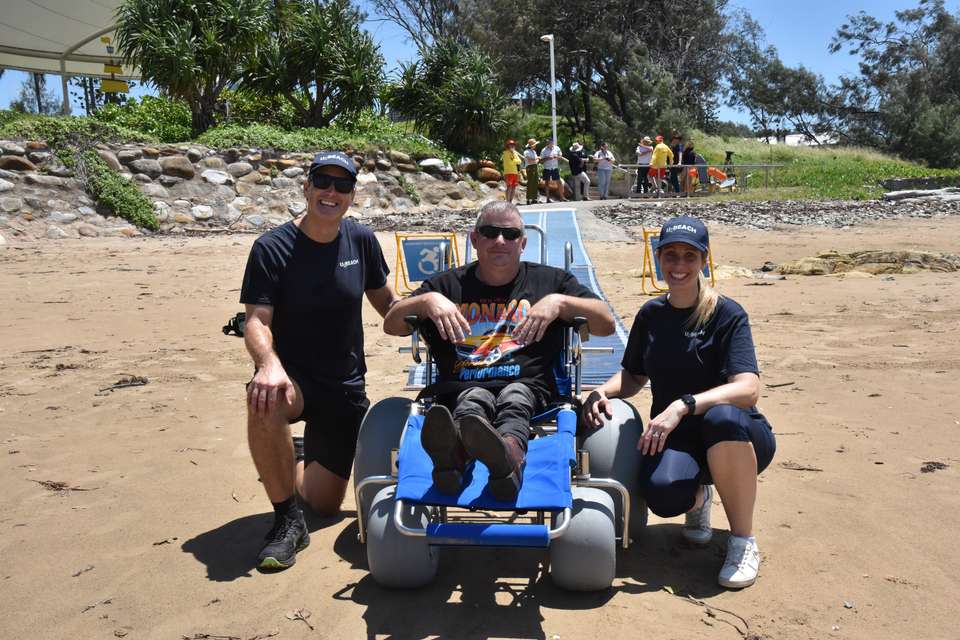 On a sandy beach, U-BEACH volunteers kneel next to a beach accessible wheelchair, while a male participant sits in the chair.