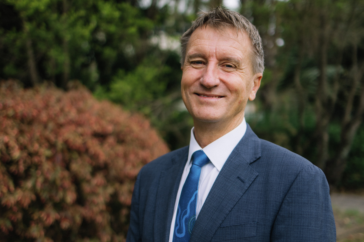 Nick Klomp wearing a navy suit, standing in front of greenery and smiling at camera with arms at his side