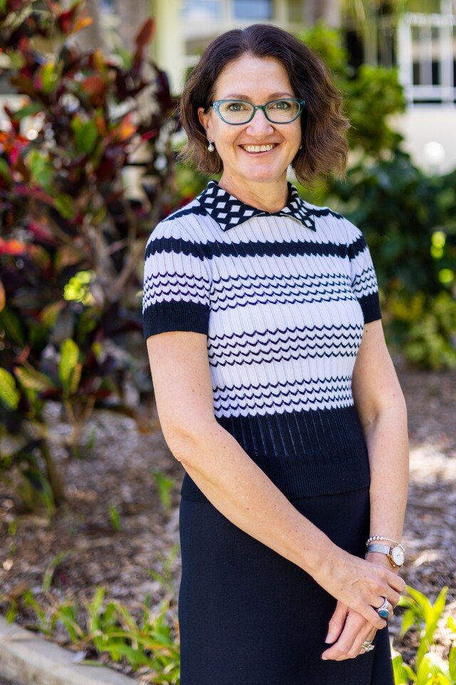 Professor Michelle Bellingan standing in front of camera smiling while wearing a white and black stripe shirt and black skirt.