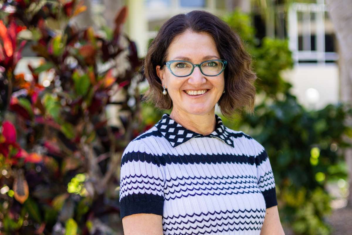 Professor Michelle Bellingan standing in front of camera smiling while wearing a white and black stripe shirt and black skirt.
