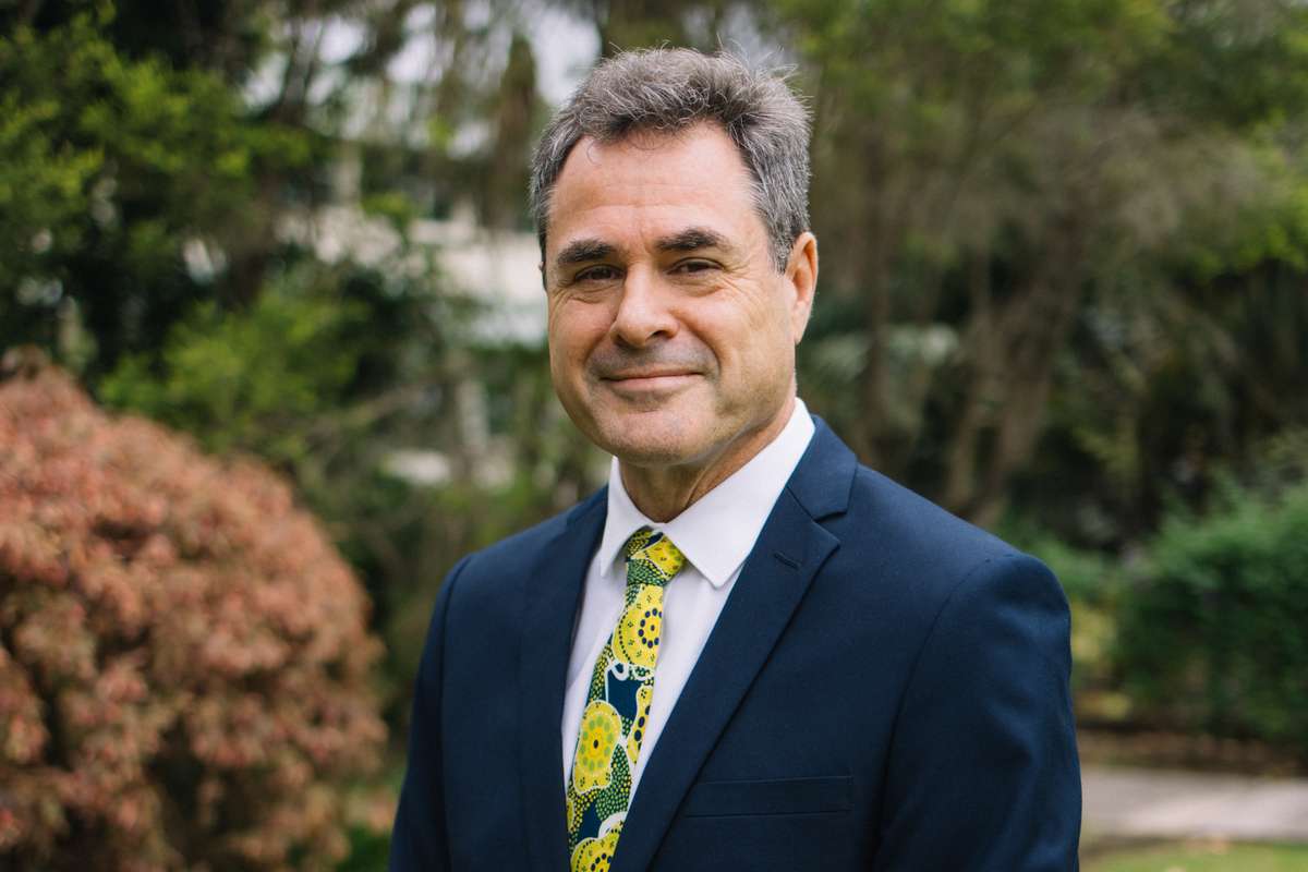 Grant Stanley in a navy suit standing in front of greenery smiling at the camera with his hands folded in front of him