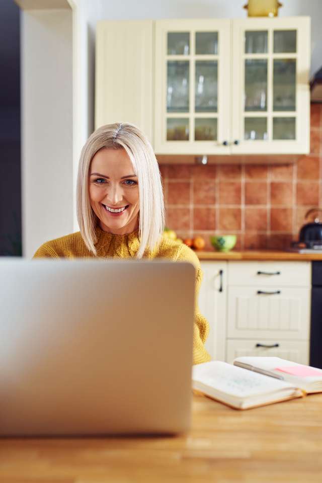 Smiling mature woman using laptop at home sitting in the kitchen.