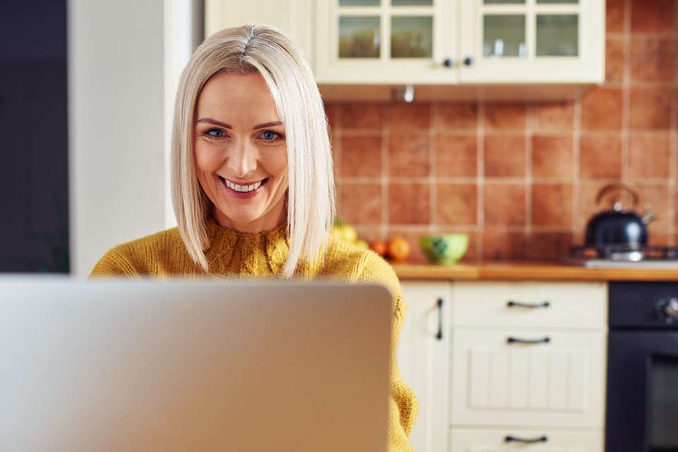 Smiling mature woman using laptop at home sitting in the kitchen.